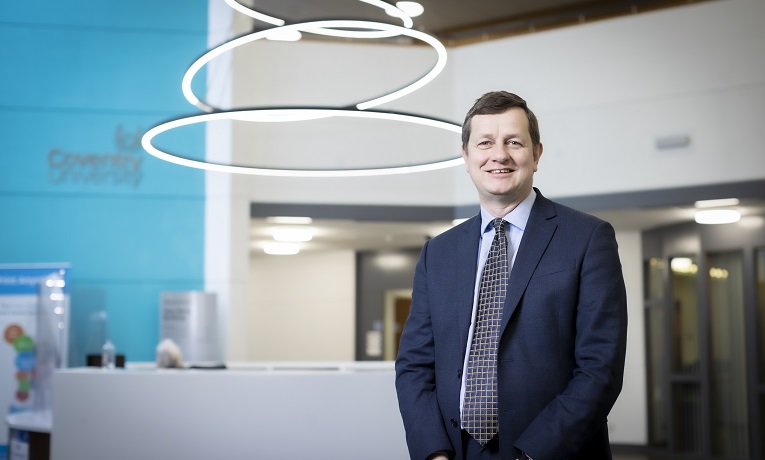 Richard Well is a suit, shirt and tie stood in a brightly lit reception area with Coventry University logo on the wall behind him