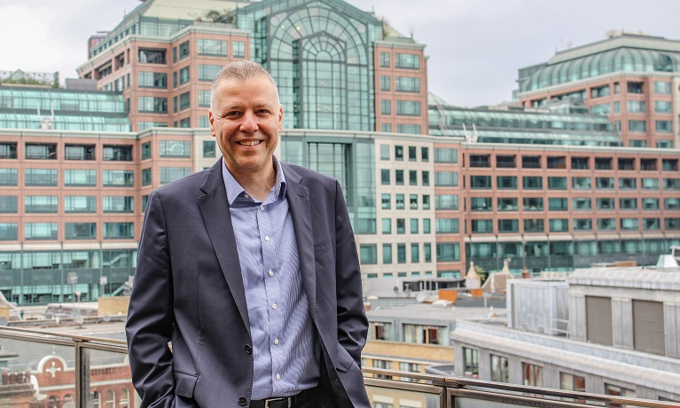 Socrates Karidis in a shirt and suit with a large brick and glass building in the background