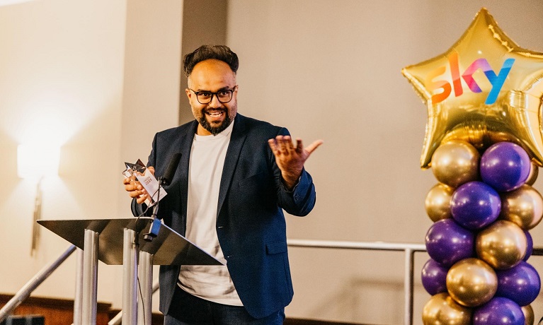 Danny Mirza collecting his award on stage with purple and gold balloons in the background
