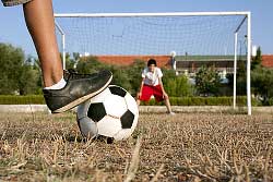 Close up of a football with a foot on it, in the background a goal keeper in a football goal