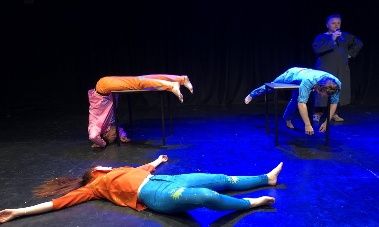 Three actors laying in different positions on a stage with a man standing in the background with a microphone