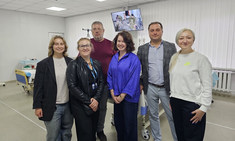 Dr Agnieszka Lewko with healthcare experts and academics stood together smiling at the camera with a hospital bed and screen in the background