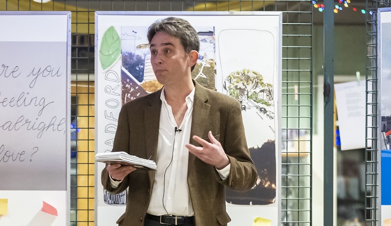 A man holding a notepad speaking in front of some framed drawings