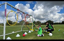 A boy holding a foam bat kneeling down on grass alongside a Coventry University researcher next to a football goal with coloured hoops attached to the crossbar