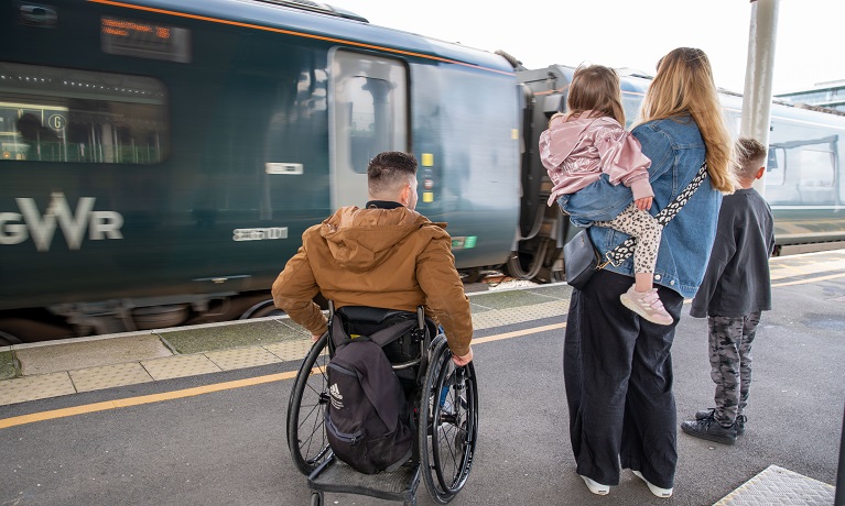 A man in a wheelchair alongside a woman holding a young girl and a boy next to them on a train station platform with a train in the background