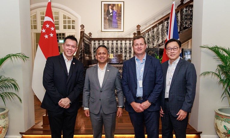 Derrick Chang, Nik Mehta OBE, Richard Wells and Michael Yap stood in a row infront of a wooden staircase and the flags of the United Kingdom and Singapore