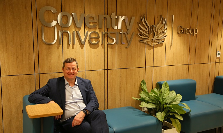 Coventry University's Deputy Vice-Chancellor (International), Richard Wells, sat in a chair wearing a suit and behind him on a wall is the Coventry University Group logo