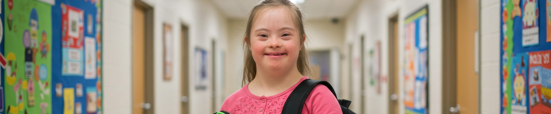 A young girl with Down syndrome smiling in a school corridor.