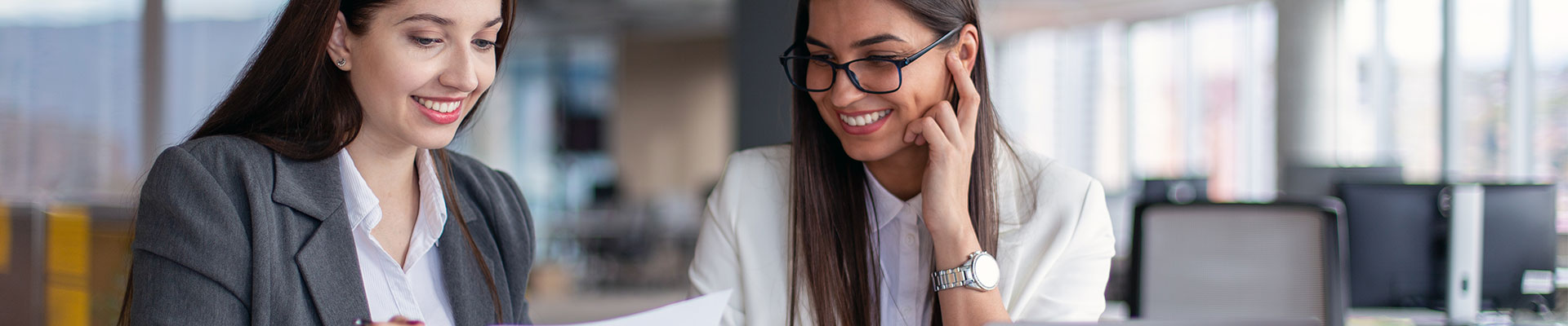 Two women in business wear looking over some paperwork at a table with a laptop open in front of them.