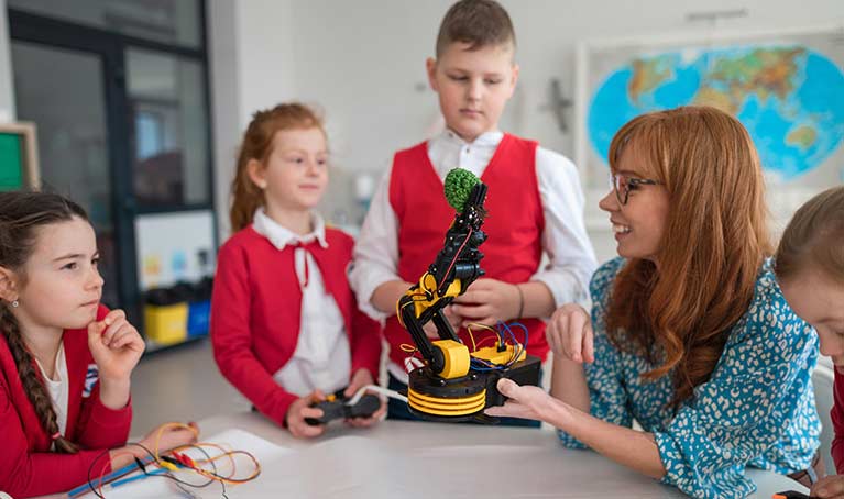 Teacher in a class room holding a tablet standing beside a girl wearing a VR headset