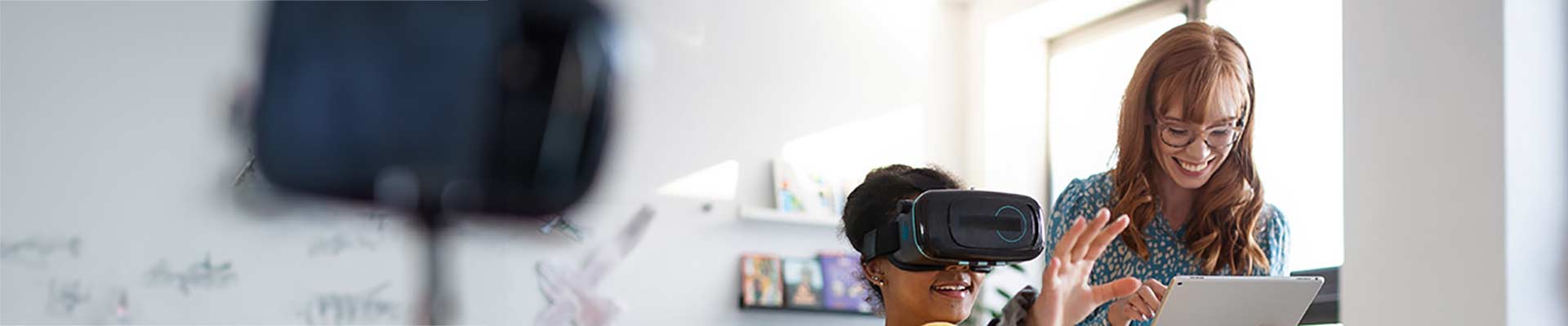 Teacher in a class room looking at a tablet standing beside a young school girl wearing a headset