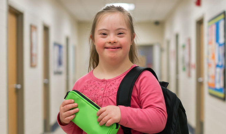 A young girl with Down syndrome smiling in a school corridor.