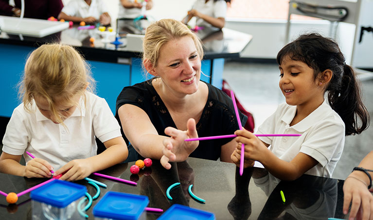 A woman teacher with blonde hair kneeling down next to a student using chemistry molecular models.