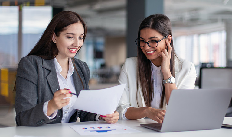 Two women in business wear looking over paperwork with a laptop open in front of them.