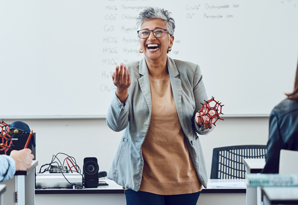 A teacher with short grey hair laughing as she teaches at the front of a classroom, holding a chemistry model.