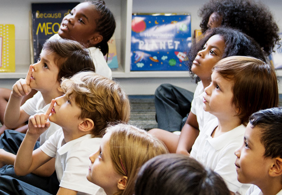 Primary schoolchildren sat on the classroom floor in a reading area looking at their teacher who is out of the shot.