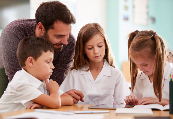 Students sitting at a table with a male teacher helping them.