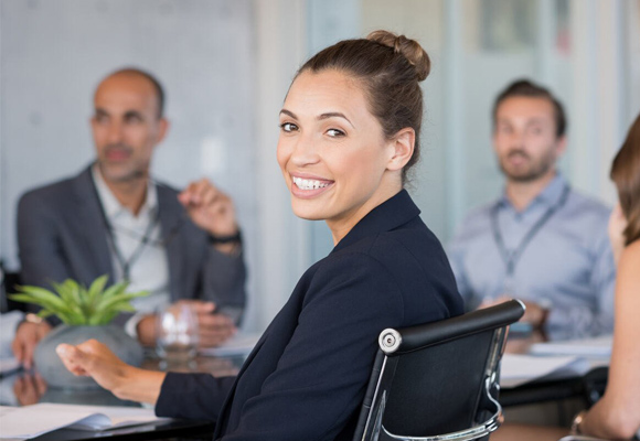 A woman looking over her shoulder to smile at the camera, sitting around a table of people in business wear.