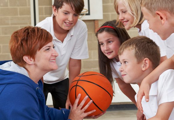 A teacher holding a basketball in a school sports hall, giving a team talk to a group of primary school-aged children.