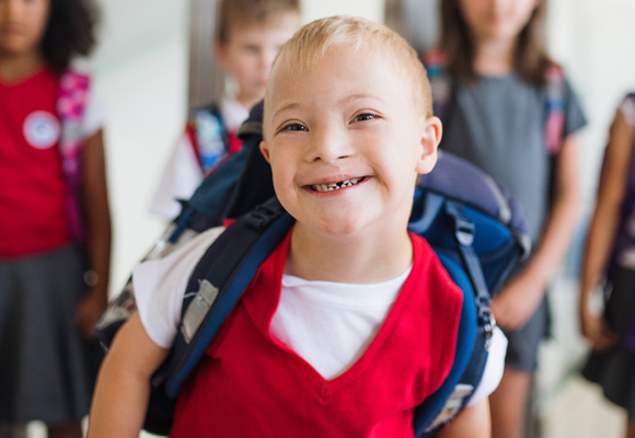 A young boy with Down syndrome smiling in a school corridor with a backpack on and students in the background behind him.