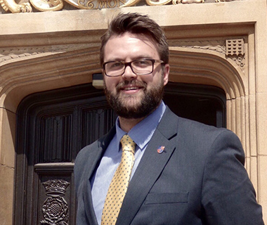 Matthew Tragheim in a suit standing in athe doorway of a historical building, smiling at the camera.