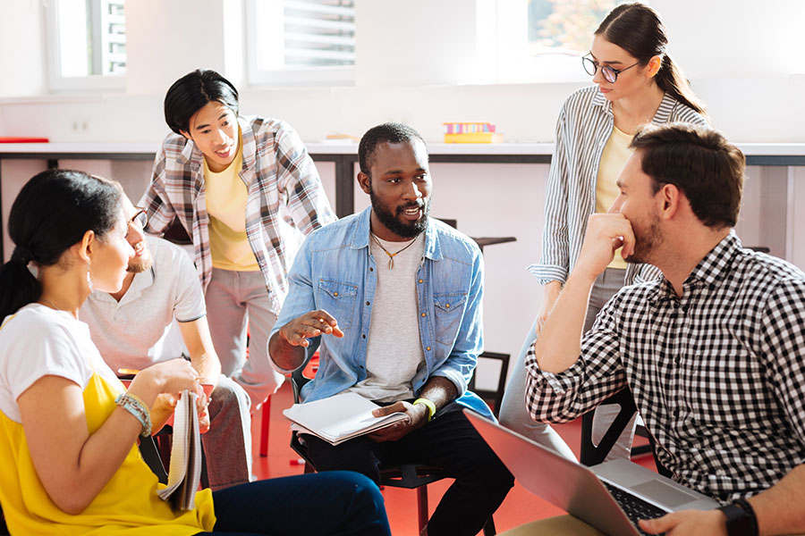 A diverse group sat having a discussion, one holding a laptop and another a clipboard.