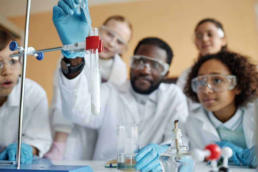 A teacher with a class of students all wearing safety glasses as he droppers liquid into a test tube.