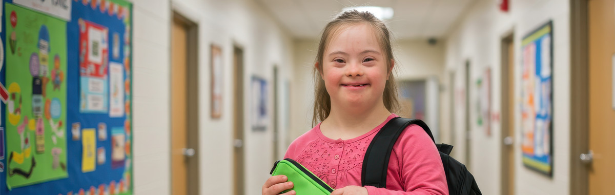 A young girl with Down syndrome smiling in a school corridor.