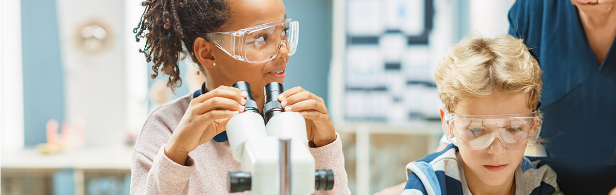 Two young students wearing goggles and using a microscope in a science classroom.