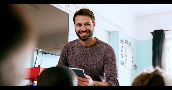 Male teacher stood at the front of a class holding a book