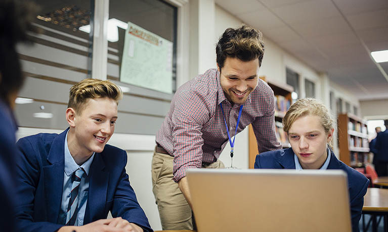 Teacher leans over one student as they work, another student to the lefthand side, all smiling