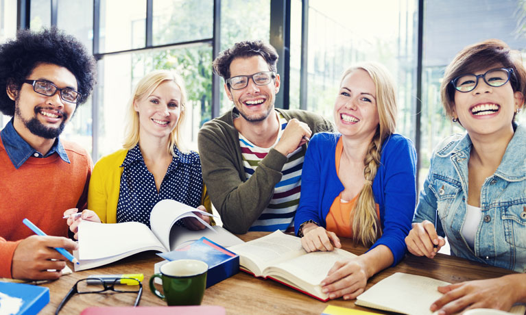 A group of people studying with papers and books in front of them, and smiling at the camera.