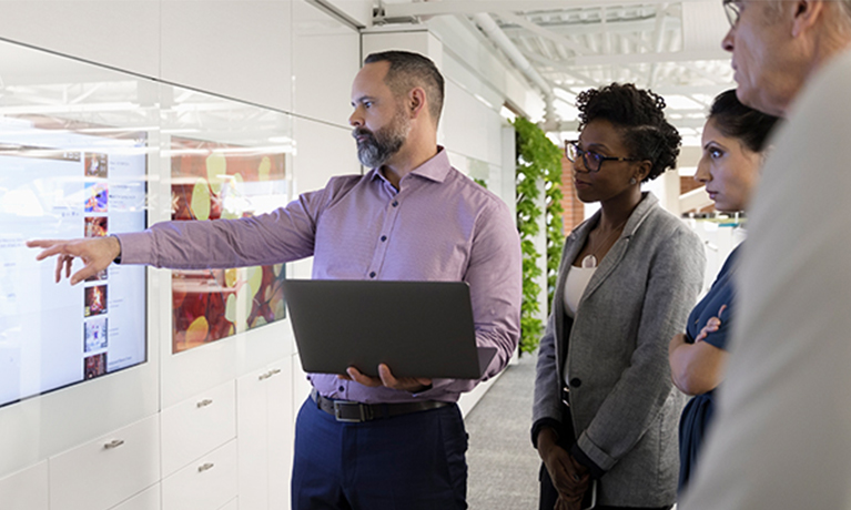 Man standing up with a laptop pointing at presentation screen to a group of people