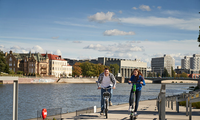 Man riding a bike and girl riding an electric scooter along the water front on a clear day.